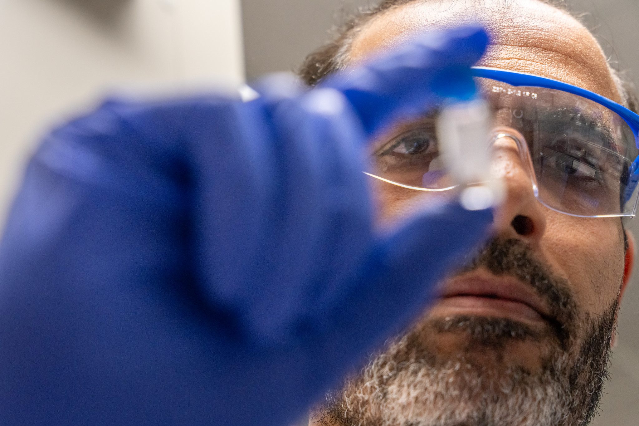 Research assistant professor Sherif Ramadan holds a small vial up to the camera. He wears a blue glove and goggles.